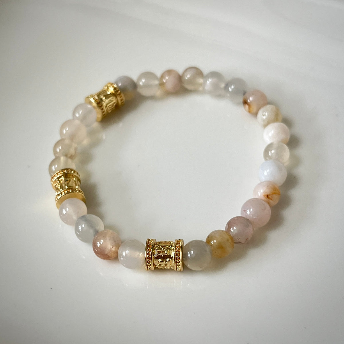 A beaded stretch bracelet featuring translucent pale pink, white, and gray gemstone beads with two ornate gold-toned cylindrical spacers, displayed on a clean white surface.